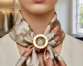 May include: A floral silk scarf in shades of olive green, cream, and rose pink, secured with a gold-tone ring. The ring is engraved with the word "CELINE PARIS". The scarf is draped around the neck.