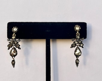 May include: A pair of silver dangle earrings with a teardrop-shaped diamond at the bottom and smaller diamonds surrounding it. The earrings are on a black display stand.