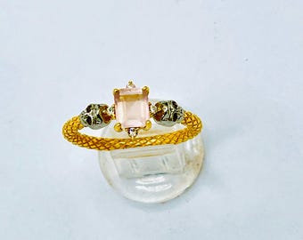 May include: Gold-toned ring with a rectangular, light pink gemstone center. The ring features a textured band and skull accents on each side of the gemstone. Small, clear stones are set near the center stone.