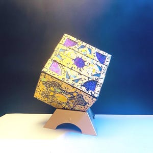 Könnte beinhalten: Ein Puzzlewürfel mit gelber Grundfläche, verziert mit komplexen schwarzen Linien und eingelegten violetten, blauen und grünen Glitzerpaneelen. Der Würfel ist schräg auf einem soliden, glatten, hellbraunen, bogenförmigen Kunststoffständer platziert.
