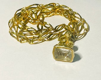 May include: A gold-colored necklace with an ornate chain design and a rectangular pendant. The pendant has a clear center with an inscription. The necklace is displayed on a white surface.