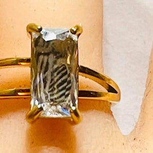 May include: A gold ring featuring a large, rectangular, clear gemstone. The ring has a double-band design, with a second band running parallel to the main band. The gemstone is set in a four-prong setting.