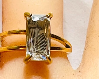 May include: A gold ring featuring a large, rectangular, clear gemstone. The ring has a double-band design, with a second band running parallel to the main band. The gemstone is set in a four-prong setting.