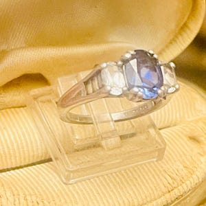 May include: A silver ring with a large, oval, dark blue gemstone as the centerpiece, flanked by two smaller, clear stones. The ring is set on a clear plastic display stand, against a soft, golden-yellow background.