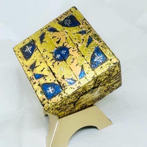 May include: A gold-colored, multi-layered cube with blue and white details sits on a gold-colored stand. The cube has a textured surface and appears to be made of metal.