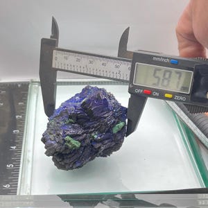 May include: A dark blue and green mineral specimen is measured with a digital caliper. The caliper displays a measurement of 58.7 mm. The mineral has a rough, textured surface and is displayed on a glass surface.