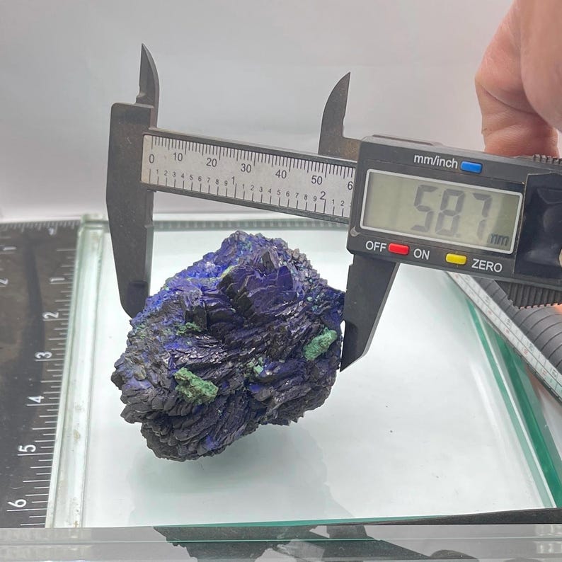 May include: A dark blue and green mineral specimen is measured with a digital caliper. The caliper displays a measurement of 58.7 mm. The mineral has a rough, textured surface and is displayed on a glass surface.
