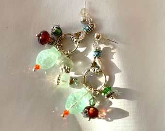 May include: Dangle earrings with silver-toned hand-shaped charms, adorned with light green, red, and clear beads. The earrings feature a mix of textures and colors, creating a unique and eye-catching accessory. The earrings are designed to be worn with pierced ears.