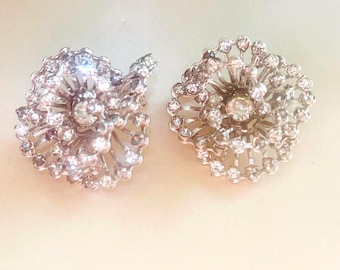 May include: Two silver-toned, flower-shaped earrings. Each earring features a central, large, clear gemstone surrounded by smaller, clear gemstones. The earrings have a detailed, openwork design, creating a sparkling, elegant appearance. The earrings are set against a soft, off-white background.