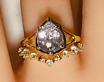 May include: A gold ring featuring a large, pear-shaped, light-colored gemstone with a dark border. The ring has a halo of smaller, round gemstones and a decorative band.