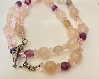 May include: A beaded necklace featuring a mix of pink, purple, and clear gemstones. The necklace has a toggle clasp and is composed of round and oval beads. The color palette includes shades of rose, lavender, and crystal.