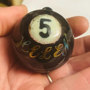 May include: A black ceramic ball with a white circle containing the number "5" and gold lettering that says "Elle" around the circle.