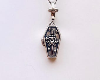May include: A silver-toned metal necklace featuring a coffin-shaped pendant. The pendant displays an embossed sword design surrounded by small star motifs on a dark, recessed background. The piece hangs from a delicate silver-toned chain link.