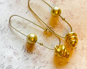 May include: A pair of gold-colored earrings. Each earring features a large, round bead and a spiral-shaped charm. The earrings are made of a thin, wire-like material and are displayed on a textured, off-white surface.