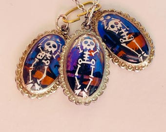 May include: A silver necklace with three oval charms featuring skeletons with colorful backgrounds. The charms have a decorative silver border.