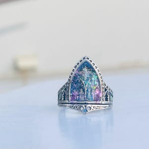 May include: A silver ring with a pointed, arched top. The top is filled with a clear resin that contains iridescent glitter and a small, white flower.