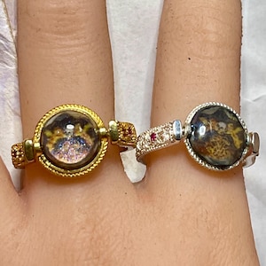 May include: Two rings worn on adjacent fingers. The left ring is gold-toned with a textured band and a circular setting containing a dark, iridescent stone. The right ring is silver-toned with a pavé-set band, a small red accent, and a dark stone.