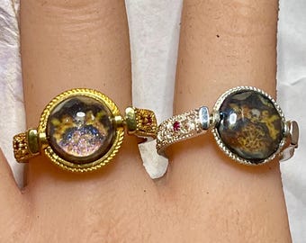 May include: Two rings worn on adjacent fingers. The left ring is gold-toned with a textured band and a circular setting containing a dark, iridescent stone. The right ring is silver-toned with a pavé-set band, a small red accent, and a dark stone.