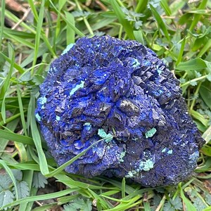 May include: A striking, natural azurite specimen. The mineral features a deep blue color with areas of black and patches of light green. The azurite is set against a backdrop of green grass.