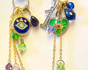 May include: Two ornate earrings with dangling charms. One features a blue eye design, while the other has a green heart. Both have gold chains and colorful beads, including green, purple, and blue. The earrings are suspended from silver hand-shaped clasps.