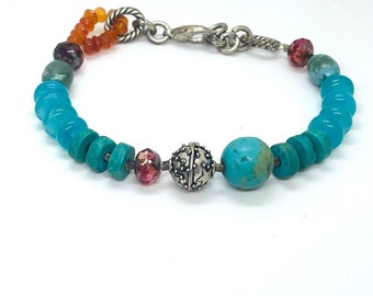 May include: A beaded bracelet featuring turquoise, teal, and orange beads. The bracelet includes a large turquoise bead, a silver bead with star designs, and a variety of smaller, colorful beads. The bracelet is connected with a silver chain and clasp.