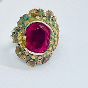 以下が含まれることがあります： テクスチャ加工されたゴールド調の金属製のセッティングに、大きな深紅の宝石がセットされたリング。セッティングは、小さな丸いアクセントで飾られています。