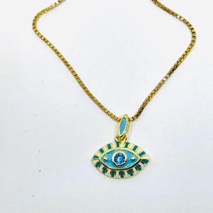 May include: Gold-toned necklace featuring an eye-shaped pendant. The pendant has a light blue enamel background with white and gold accents, and a small blue gemstone in the center. The chain is a delicate, box-style link.