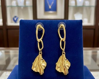 May include: A pair of gold-toned drop earrings featuring an irregular, organic-shaped pendant suspended from two interlocking, uneven oval links. The earrings are displayed on a dark blue velvet stand, showcasing a textured, rippled surface finish.