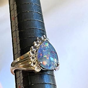 May include: A gold-toned ring featuring a large, pear-shaped opal with iridescent blue and green flashes. The opal is accented by a cluster of small, clear gemstones on one side. The ring is displayed on a black ring sizer.