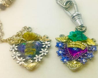 May include: Two ornate heart-shaped pendants with floral and gold accents. One pendant has white flowers and a blue heart, the other has blue flowers and a purple heart with the text "In Memory of". Both are on chains.