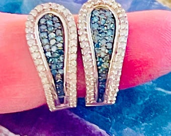 May include: A pair of silver-toned earrings with a curved, elongated design. The earrings feature a central cluster of small, blue-green gemstones surrounded by a border of clear, sparkling stones. The earrings are set against a pink and purple background.