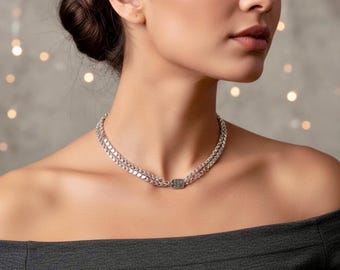 May include: A double-strand silver chain necklace featuring a small, rectangular, textured metallic clasp at the center. The necklace rests closely around the neck, showcasing a polished finish and a uniform link design that connects to the central piece.