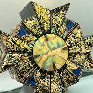 May include: A geometric puzzle box with a starburst design. The box features gold filigree, black accents, and blue and yellow iridescent details. The central circle has a marbled appearance.