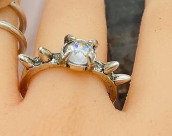 May include: A silver ring with a central, clear, faceted gemstone. The ring has a unique design with small, pointed embellishments around the gemstone and on the band. The ring is on a finger.