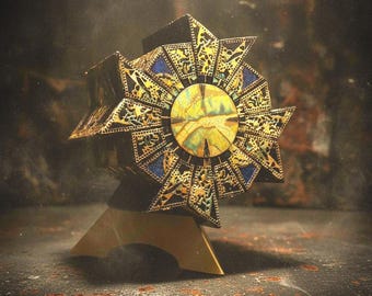 May include: A multi-faceted, star-shaped decorative object featuring intricate gold-toned filigree patterns over dark blue panels. A circular, textured yellow and green center piece sits at the core of the star, all mounted on a solid gold-toned base.