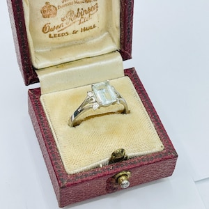 May include: A silver ring with a rectangular, light blue gemstone and small accent stones, displayed in a vintage red jewelry box with the text "Owen Robinson Ltd, Leeds & Hull."