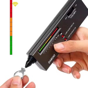 May include: A black handheld diamond tester device with a pointed probe tip touching a silver ring with a clear gemstone. The device features a volume dial, indicator lights labeled LAMP ON BATTERY OK and LAMP ON READY OK, and a vertical scale.