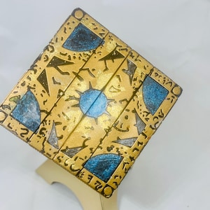 May include: A gold and blue puzzle box with intricate designs and symbols. The box is made of wood and has a layered design.
