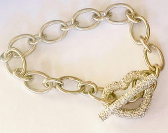 May include: A silver-toned chain bracelet with oval links. The clasp is a bar and loop design, encrusted with small, sparkling crystals. The bracelet is displayed on a white surface.