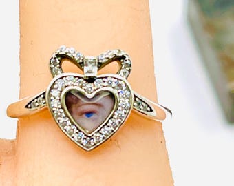 May include: Silver heart-shaped ring with a bow design at the top. The heart is encrusted with small, clear stones and surrounds a small image of a blue eye. The ring is on a finger.