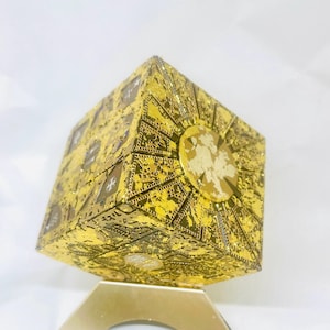 May include: A gold-colored puzzle box featuring intricate, etched geometric patterns and symbols across its six faces. The cube displays a metallic, reflective finish with detailed engravings, including circular motifs and star-like shapes on each side.