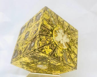 May include: A gold-colored puzzle box featuring intricate, etched geometric patterns and symbols across its six faces. The cube displays a metallic, reflective finish with detailed engravings, including circular motifs and star-like shapes on each side.