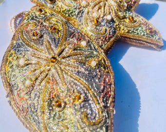 May include: Close-up of two ornate, star-shaped and teardrop-shaped ornaments. The ornaments feature intricate embroidery in gold, silver, and various colors, embellished with beads. The designs have a vintage, handcrafted aesthetic, suitable for holiday decor.