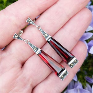May include: A pair of silver-toned drop earrings with rectangular, deep red gemstones. The earrings have a geometric design with small, decorative accents. The earrings are shown against a soft background.