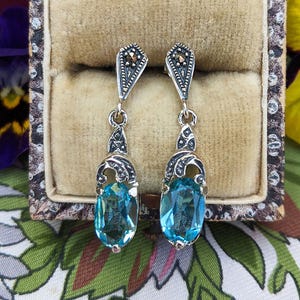 May include: A pair of silver-toned drop earrings with oval, light blue gemstones. The earrings have ornate filigree detailing and small, dark accents at the top. The earrings are displayed in a vintage jewelry box.