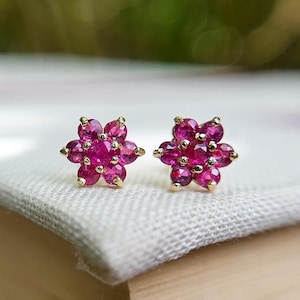 May include: A pair of stud earrings featuring a flower-shaped design. Each earring is crafted with seven round, faceted, deep pink gemstones set in a gold-toned metal setting. The stones are arranged with one central gem surrounded by six petals.