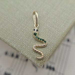 May include: A gold snake pendant featuring a series of small, round, green gemstones embedded along the upper body and head. The snake has two dark, circular eyes and a textured, segmented tail that curves into a gentle S-shape at the bottom.