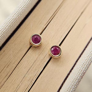 May include: A pair of round stud earrings featuring deep red, faceted circular stones set in polished gold-toned metal bezels. The earrings are positioned on a light-coloured surface with visible parallel lines, highlighting the smooth metal finish.