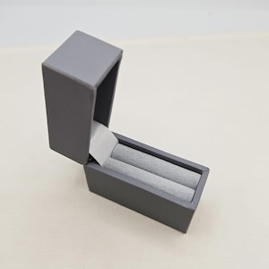 May include: A grey ring box with two compartments for rings. The box is open and the compartments are lined with grey velvet.