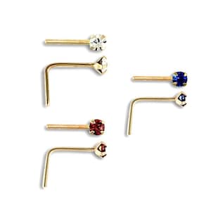 Pode incluir: Conjunto de quatro argolas de nariz de ouro com pedras preciosas de diferentes cores. As argolas são em forma de L e têm uma pequena pedra preciosa na extremidade. As pedras preciosas são transparentes, vermelhas, azuis e brancas.